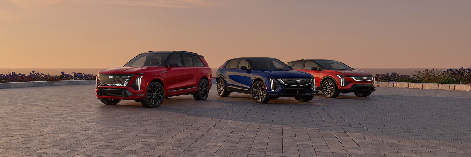 A Red Cadillac Escalade IQ, a Blue Cadillac Lyriq, and an Orange Cadillac Optiq Are Parked on a Paved Overlook with an Ocean Sunset