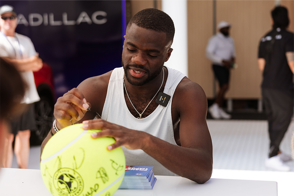 Frances Tiafoe Signing a Large Yellow Tennis Ball on a Table with Cadillac Branding in the Background