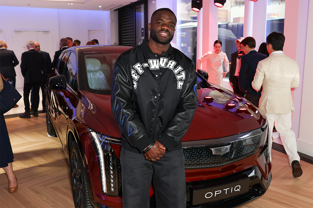 Frances Tiafoe Smiling and Standing in Front of a Dark Red Cadillac OPTIQ Electric SUV Wearing an OFF-WHITE Jacket
