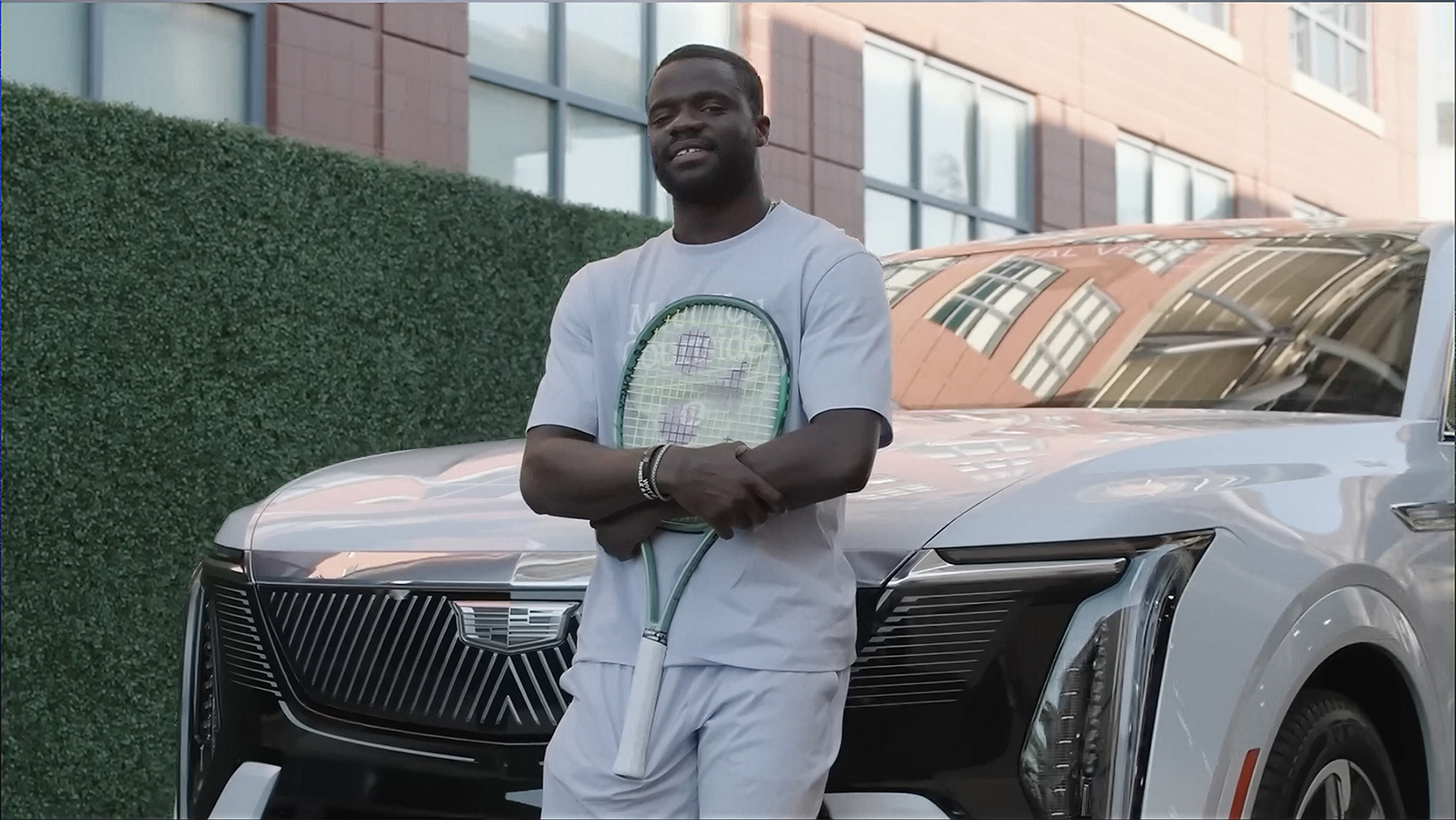 Frances Tiafoe Standing in Front of a White Cadillac SUV Holding a Tennis Racket with a Green Wall Behind Him