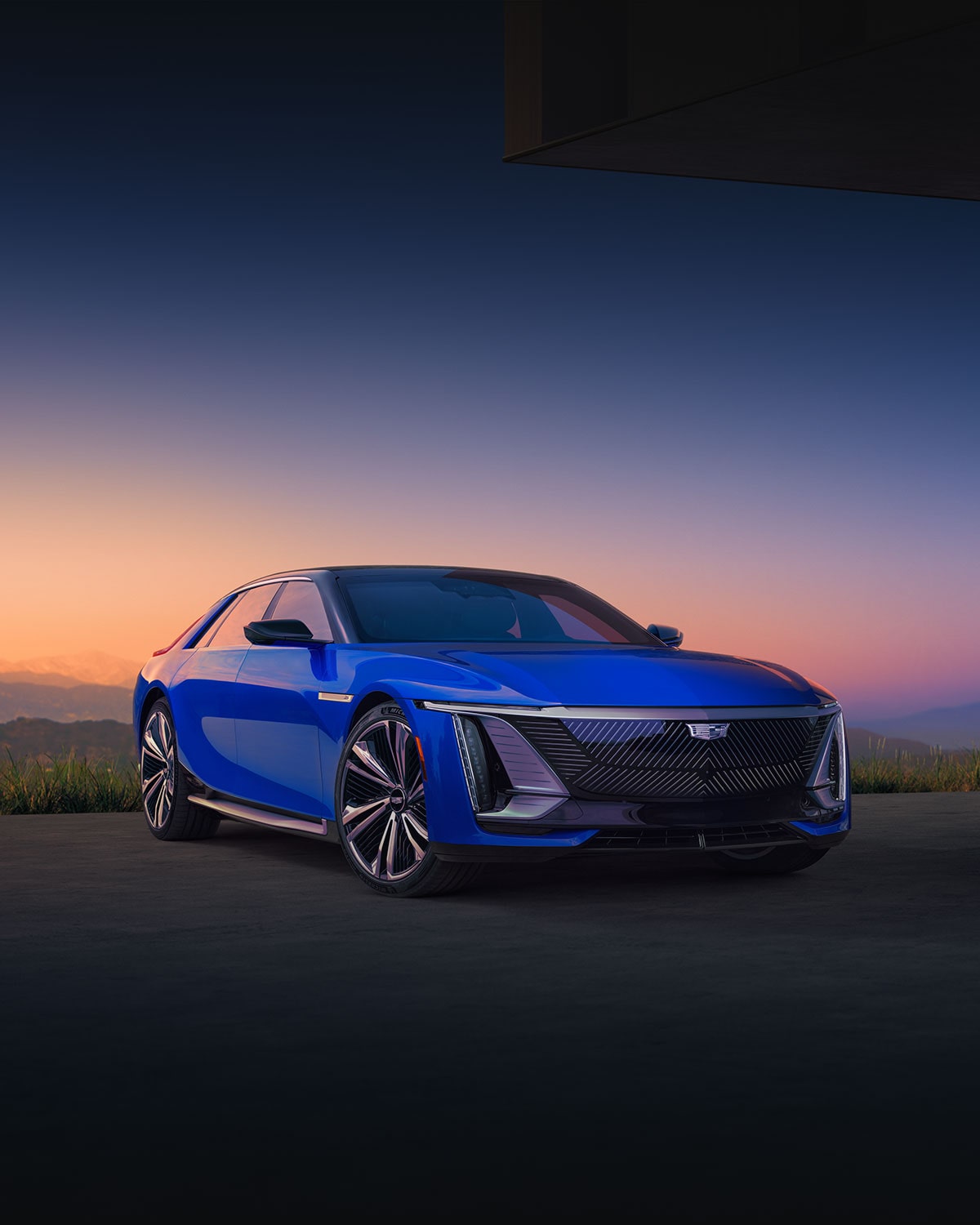 A Blue Cadillac CELESTIQ Luxury Sedan Parked Outdoors at Sunset With Illuminated Vertical LED Lighting, a Black Grille, and Mountainous Terrain in the Background.