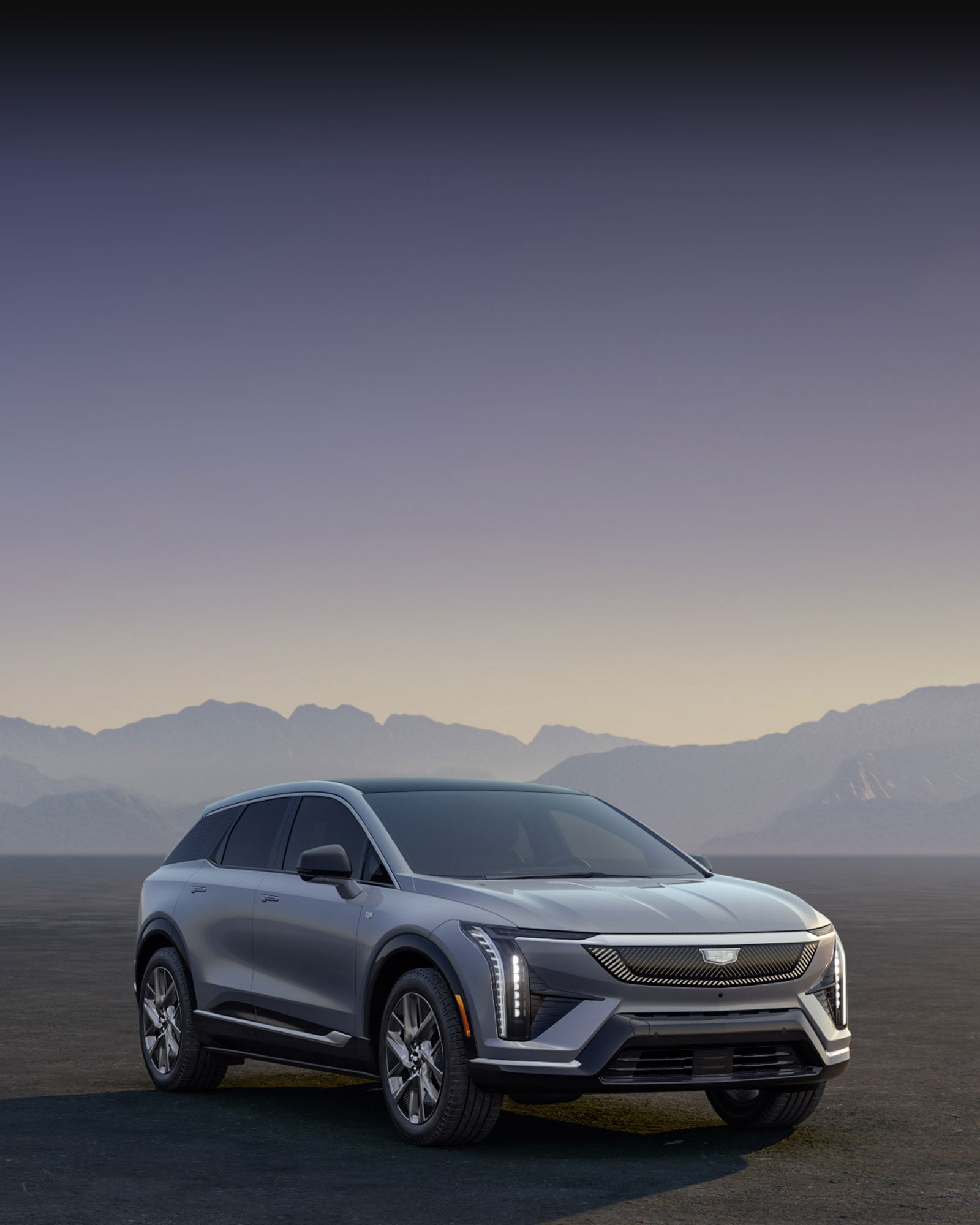 A Silver Cadillac OPTIQ Electric SUV Parked in a Desert Landscape Against a Mountain Backdrop at Dusk