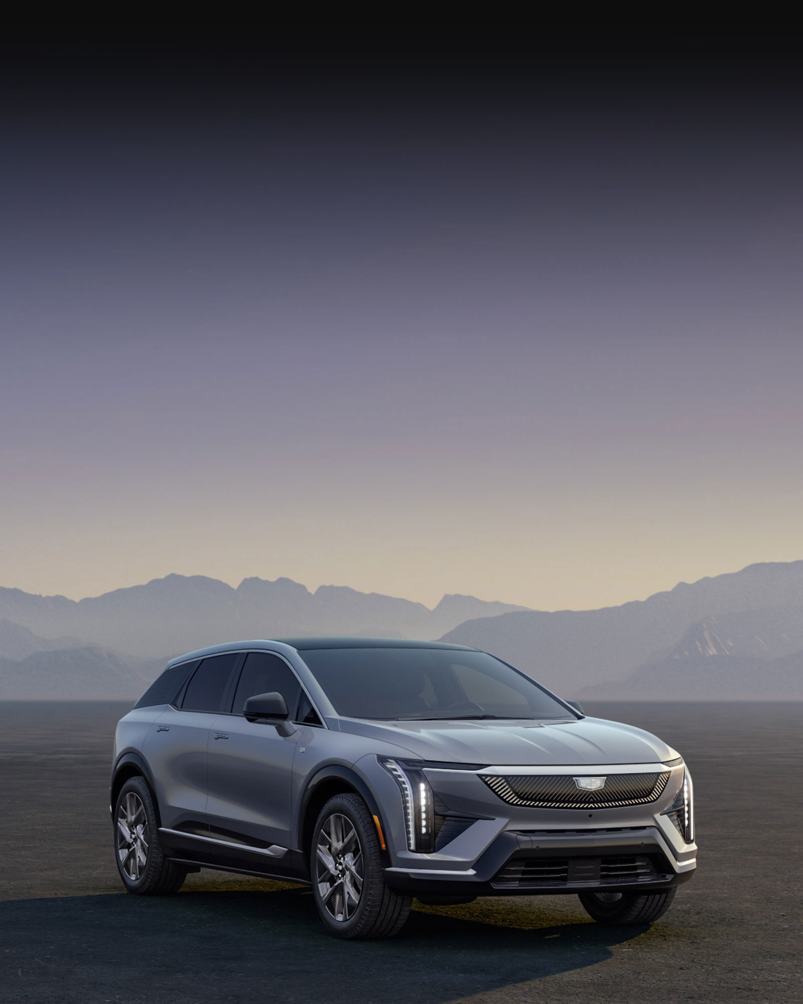 A Silver Cadillac OPTIQ Electric SUV Parked in a Desert Landscape Against a Mountain Backdrop at Dusk