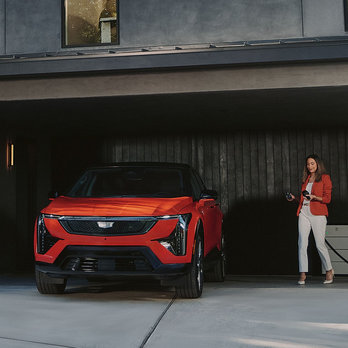 A Woman Walking Towards a Red Cadillac OPTIQ Parked in a Garage with an EV Charger in Hand