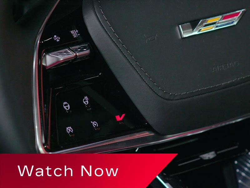 A Close-Up View Of A Cadillac OPTIQ Steering Wheel Featuring Illuminated Touch Controls And A Prominent V-Series Badge Above A Red Banner Reading “Watch Now.”
