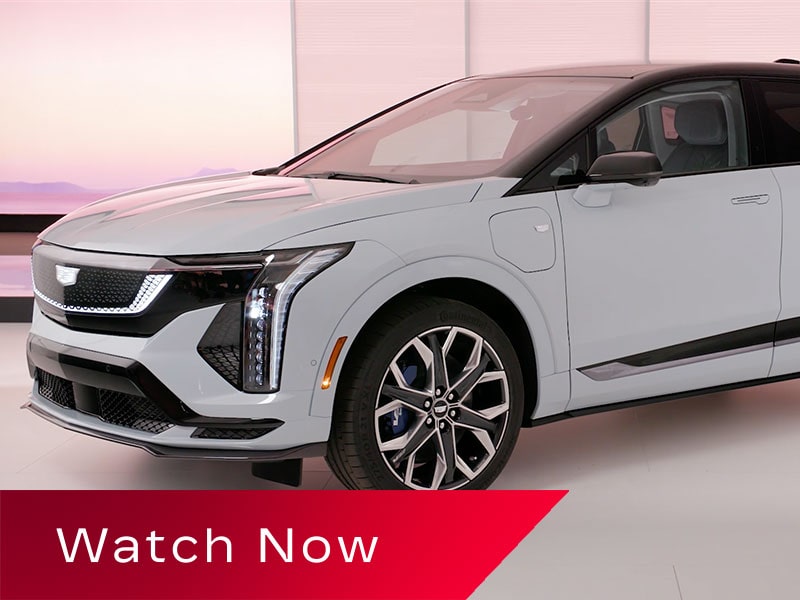 A White Cadillac OPTIQ Positioned in a Bright Studio Setting With Distinctive Vertical LED Headlamps and Multi-Spoke Wheels Above a Red Banner Reading “Watch Now.”