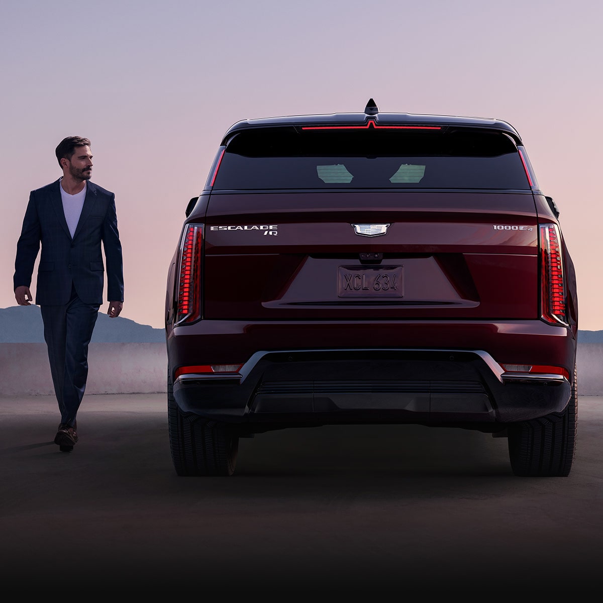 A Man Walking Away from the Rear of a Dark Red Cadillac SUV with Bold Vertical Taillights Mountains in the Distance