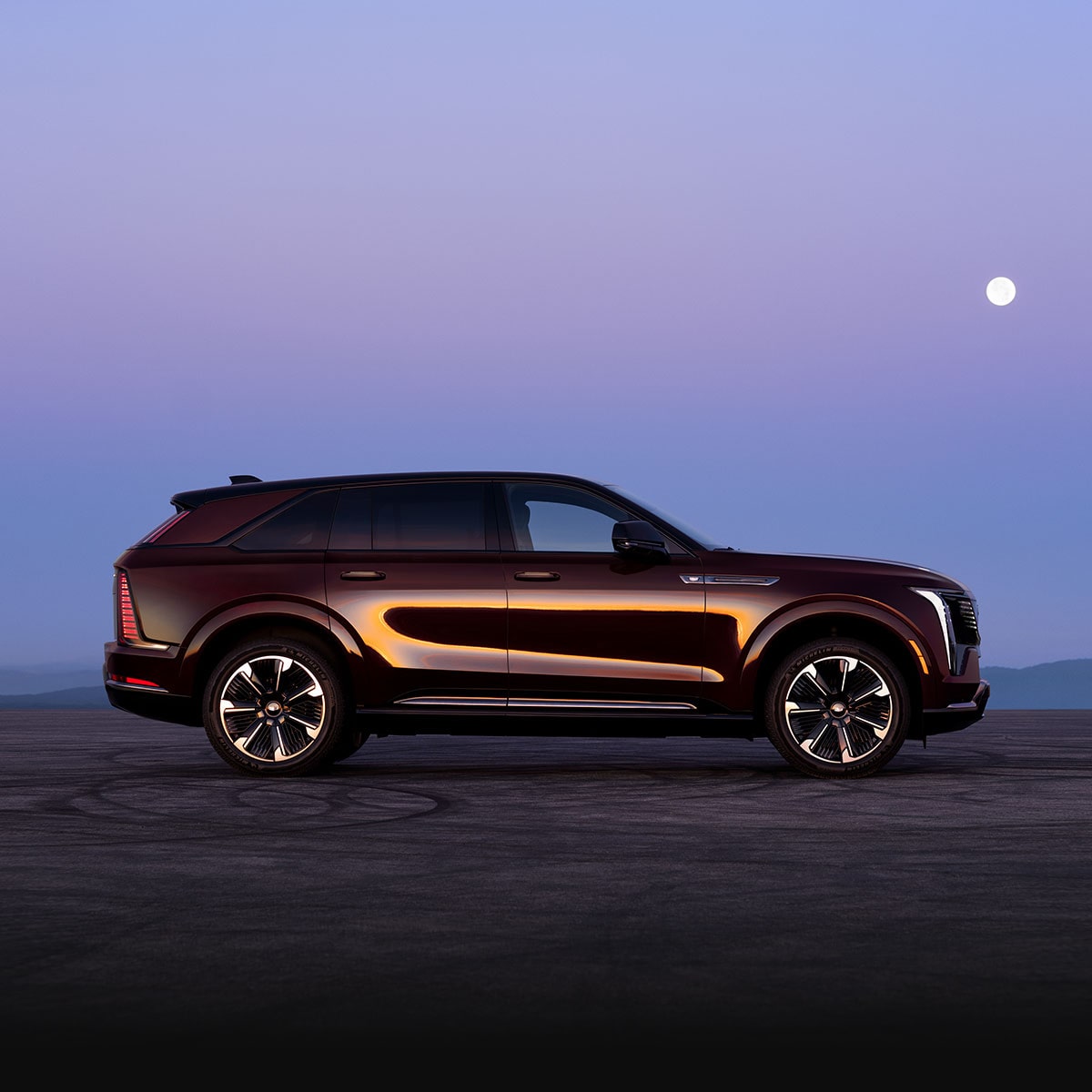 A Side Profile of a Dark Red Cadillac SUV Parked on an Open Road at Dusk with Mountains in the Background and a Bright Moon