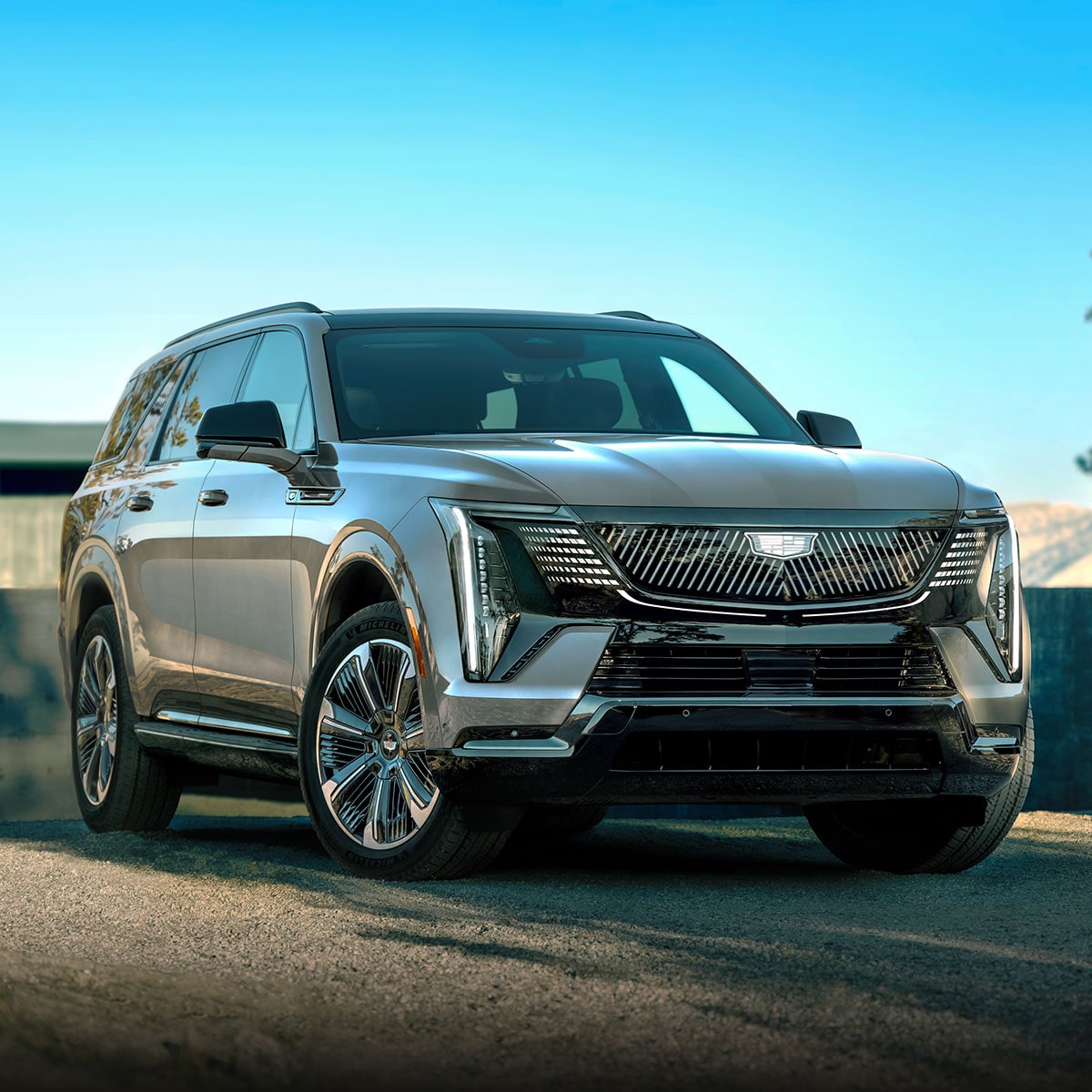 A Front Passenger Side View of a Light Silver Cadillac SUV Parked outside a Modern Building with Trees