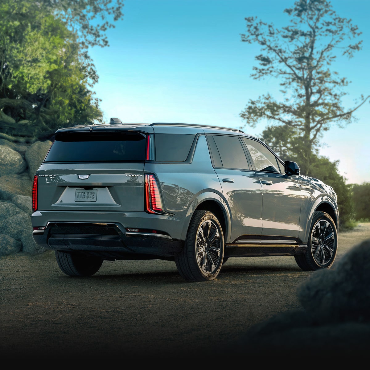 A Rear Passenger Side View of a Dark Gray Cadillac SUV Parked on a Gravel Path Surrounded by Rocks and Trees