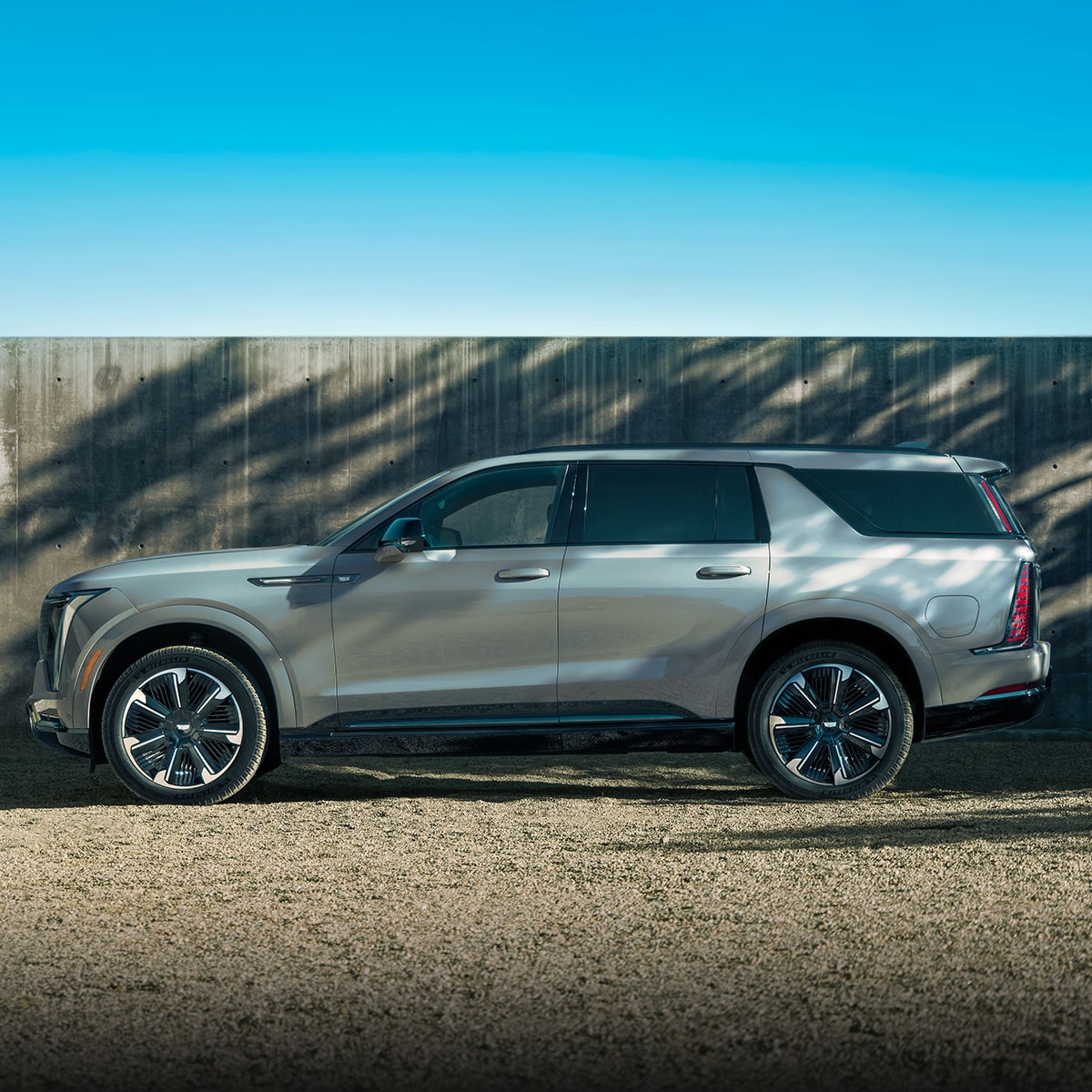 A Side Profile of a Light Silver Cadillac SUV Parked in Front of a Concrete Wall with Dramatic Shadows