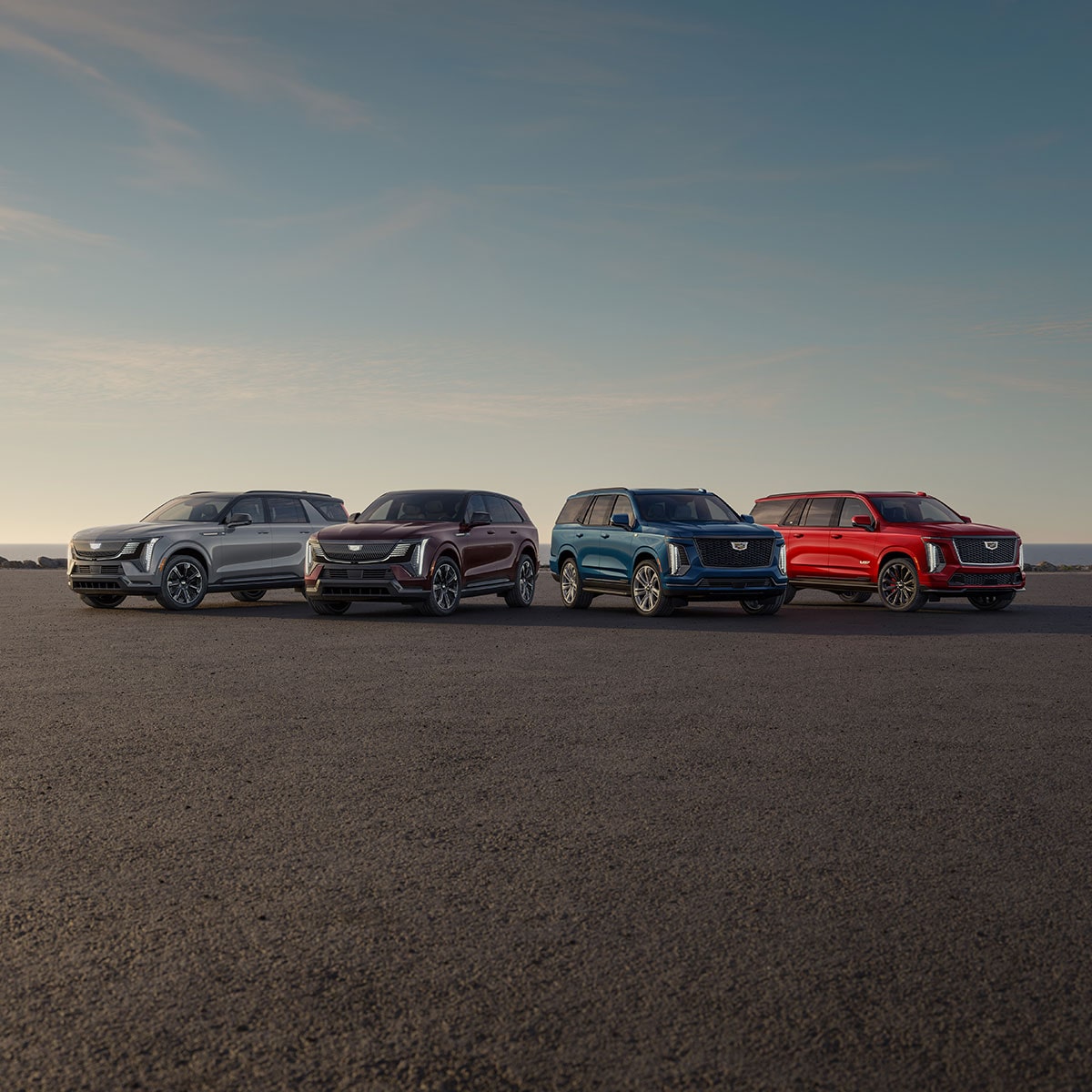 Four Cadillac SUVs, a Gray, a Maroon, a Teal Blue, and a Red Model, Are Parked on a Paved Area Facing the Ocean under a Clear Sky