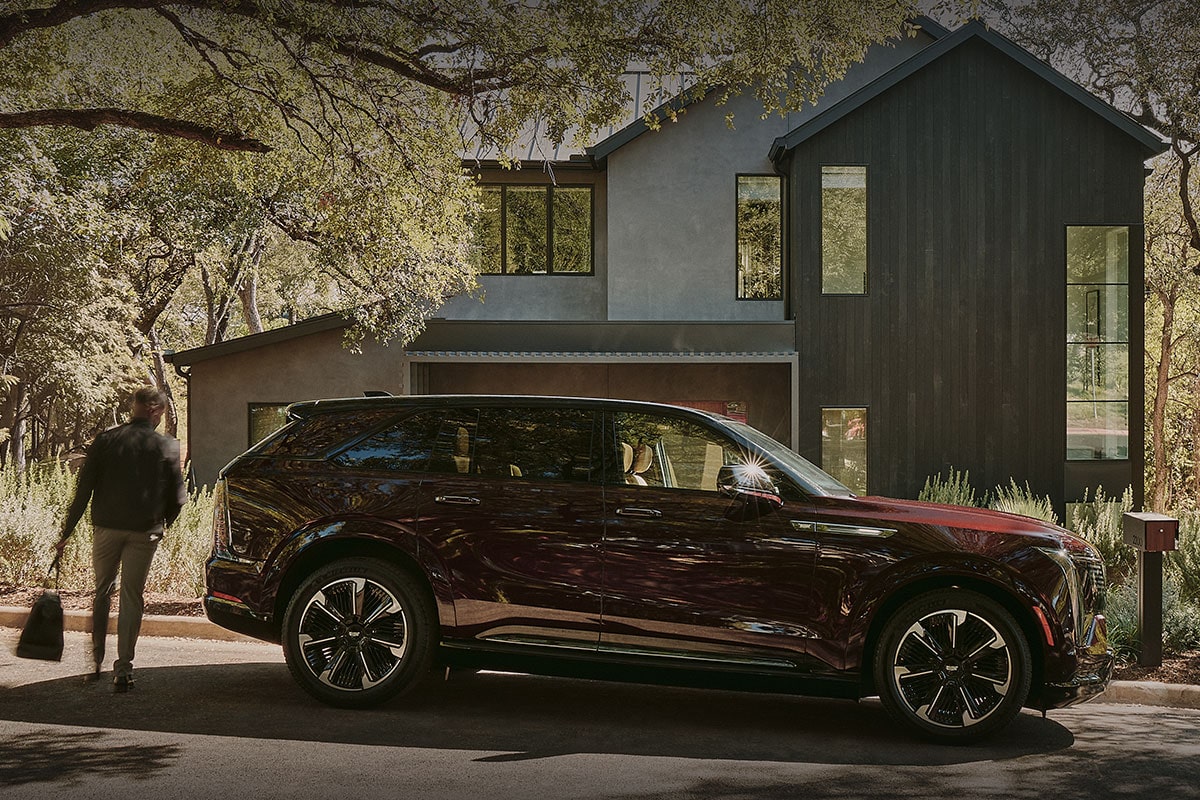 A Man with a Briefcase Walking Around the Rear of a Cadillac ESCALADE IQ Towards a Modern Home Surrounded by Trees