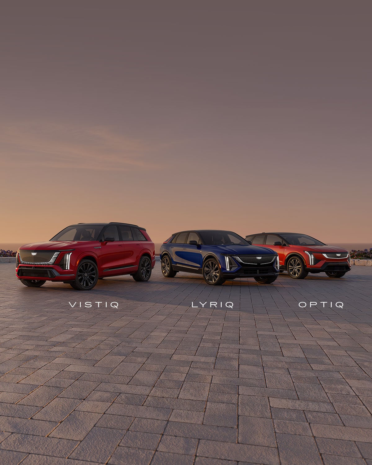 A Gray Cadillac Escalade IQ, a Red Cadillac Vistiq, and A Blue Cadillac OPTIQ Lined Up In A Sunset Background