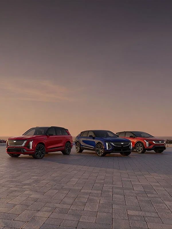 A multi-vehicle lineup of the Cadillac VISTIQ, LYRIQ, and OPTIQ parked on a paved stone driveway in front of a sunset