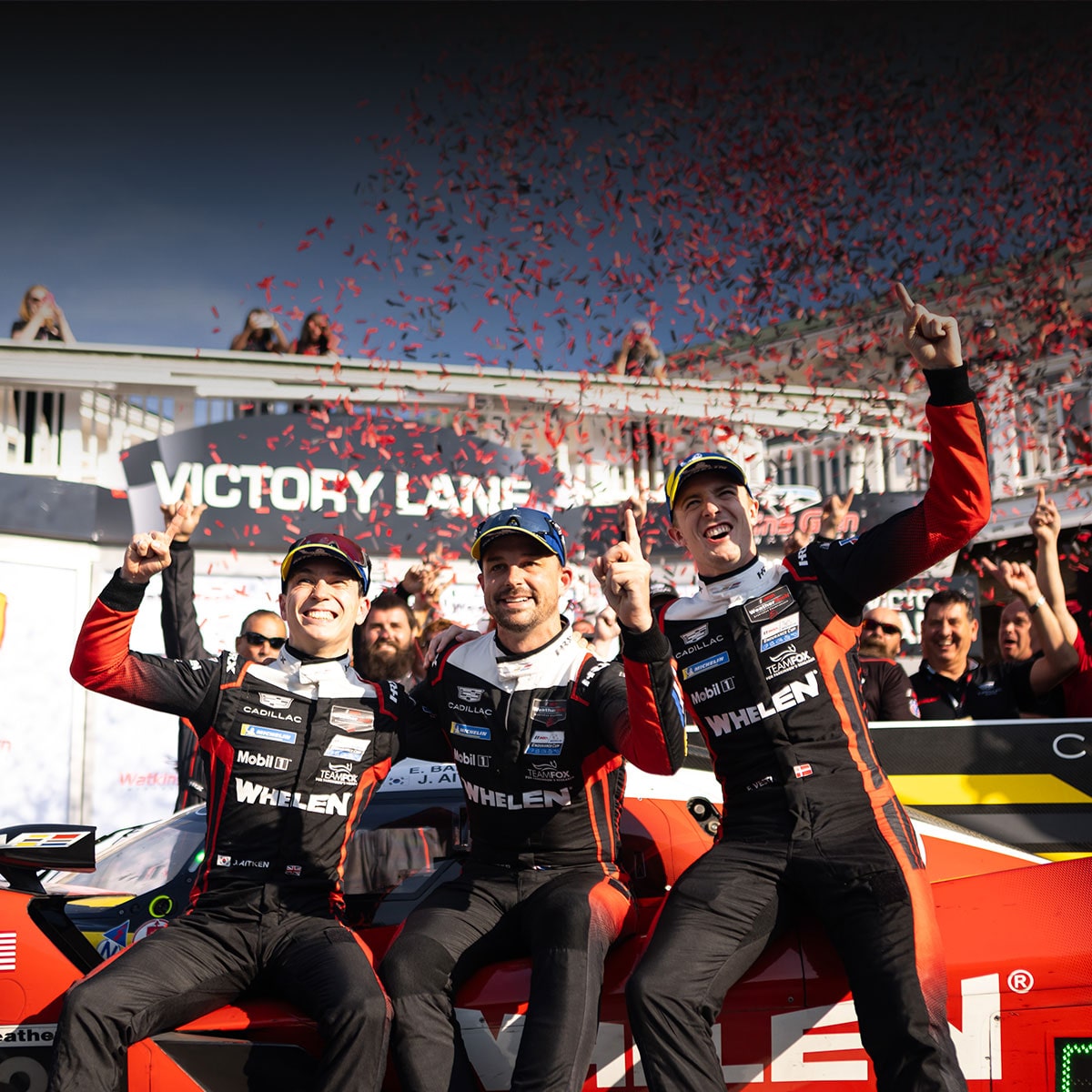 Three Cadillac Racing Team Drivers in Black Whelen-Branded Performance Suits Celebrating on a Red Cadillac Race Car in Front of a Victory Lane Sign With Red Confetti Falling Overhead.