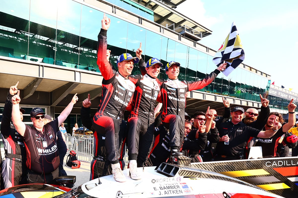 Side View of the Cadillac Racing Team at the Race Track Holding Up Their Hands and a Flag