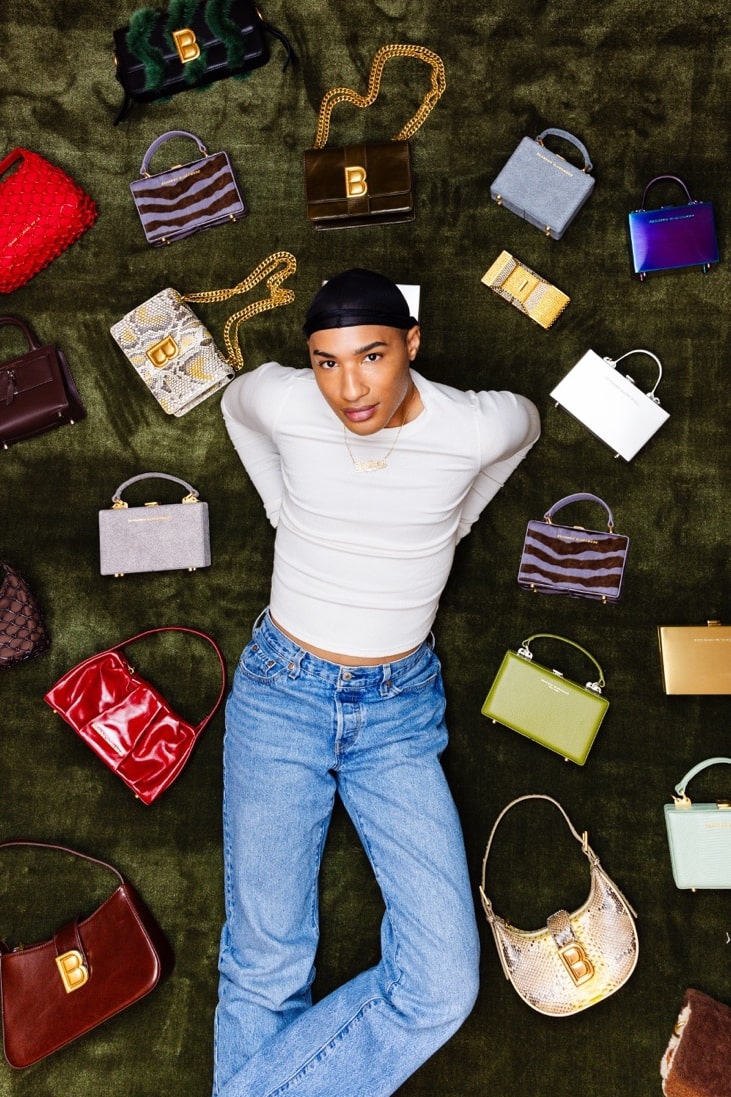 Designer Brandon Blackwood Posing and Smiling on a Dark Green Velvet Background Surrounded by Beautiful, Custom Handbags