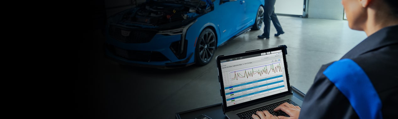 Cadillac Certified Service Technician Working on a Laptop with a Vehicle Nearby