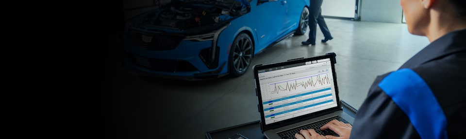 Cadillac Certified Service Technician Working on a Laptop with a Vehicle Nearby