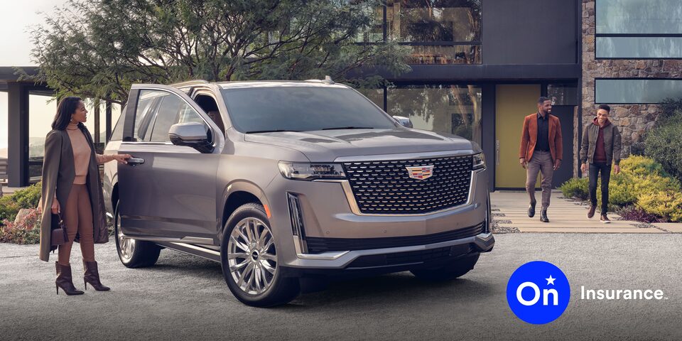 Una Cadillac Escalade en la entrada de garaje de una casa moderna. Una mujer parada al lado de la puerta abierta del lado del pasajero, mientras que un hombre y un niño salen de una casa y se dirigen hacia el auto. Logo de Seguro OnStar en la esquina inferior derecha.