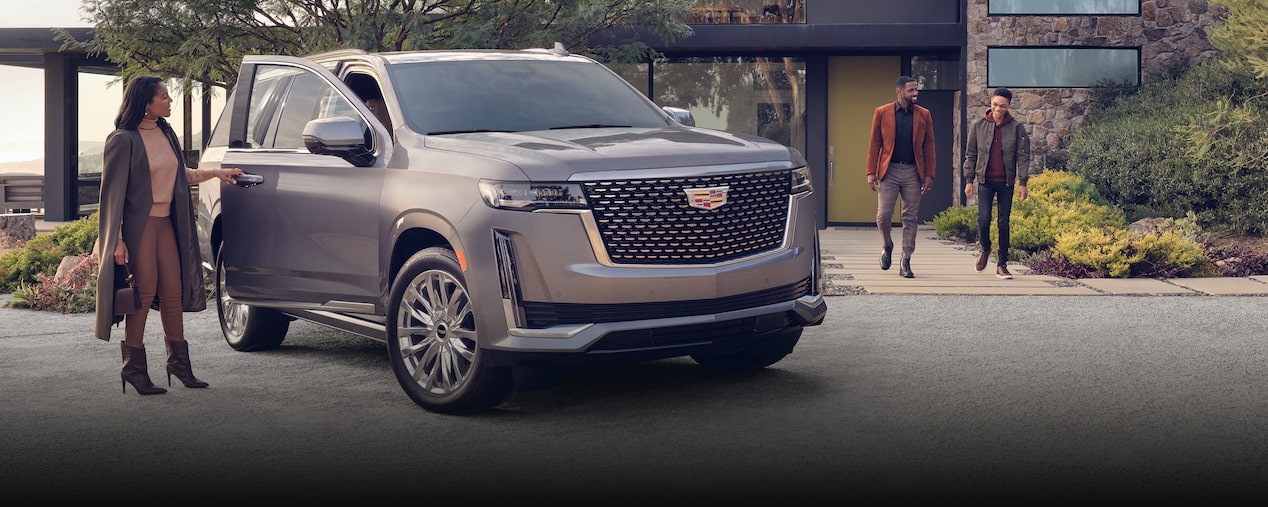 Una Cadillac Escalade en la entrada de garaje de una casa moderna. Una mujer parada al lado de la puerta abierta del lado del pasajero, mientras que un hombre y un niño salen de una casa y se dirigen hacia el auto. Logo de Seguro OnStar en la esquina inferior derecha.