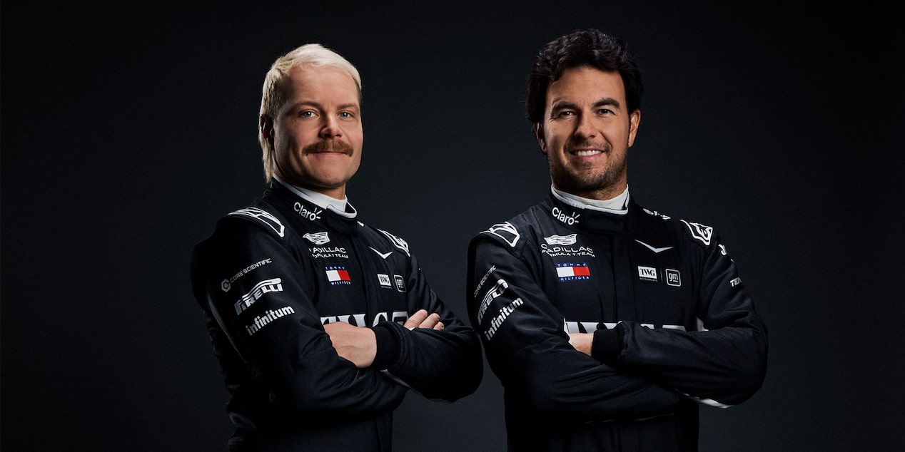 Two Team Members In Black Cadillac Racing Suits Stand Shoulder To Shoulder Against A Dark Studio Background.