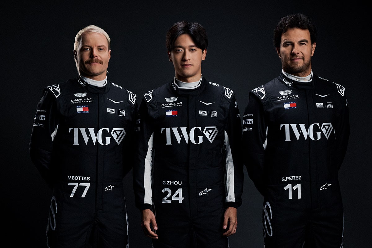 Three Team Members Wearing Matching Black Racing Suits With Cadillac And Sponsor Logos Stand Side By Side Against A Dark Studio Background.