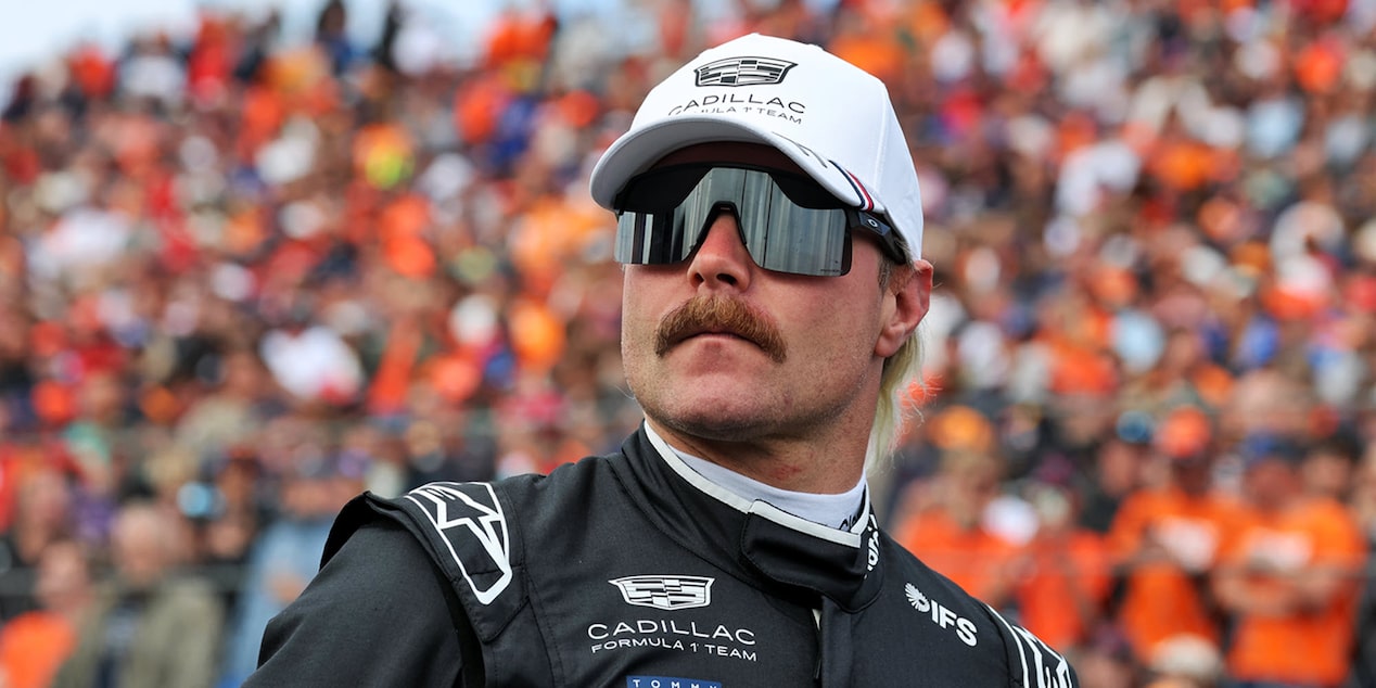 Valtteri Bottas con una gorra blanca del equipo Cadillac Formula 1, de pie en la parrilla antes de la sesión de clasificación, con anteojos de sol y mirando a lo lejos. Una multitud vestida de naranja aparece borrosa detrás de él.