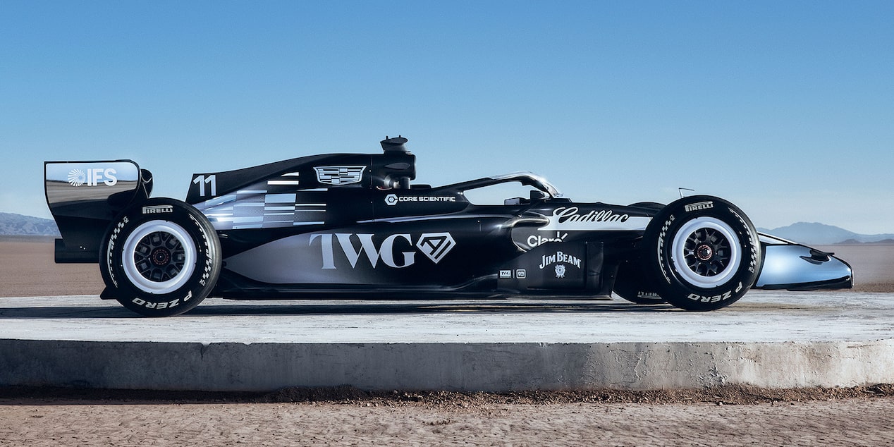 A Side Profile Of A Black-And-White Formula Race Car With Cadillac Branding Parked On A Sunlit Desert Flat With Mountains In the Background.