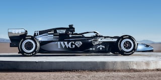 A Side Profile Of A Black-And-White Formula Race Car With Cadillac Branding Parked On A Sunlit Desert Flat With Mountains In the Background.