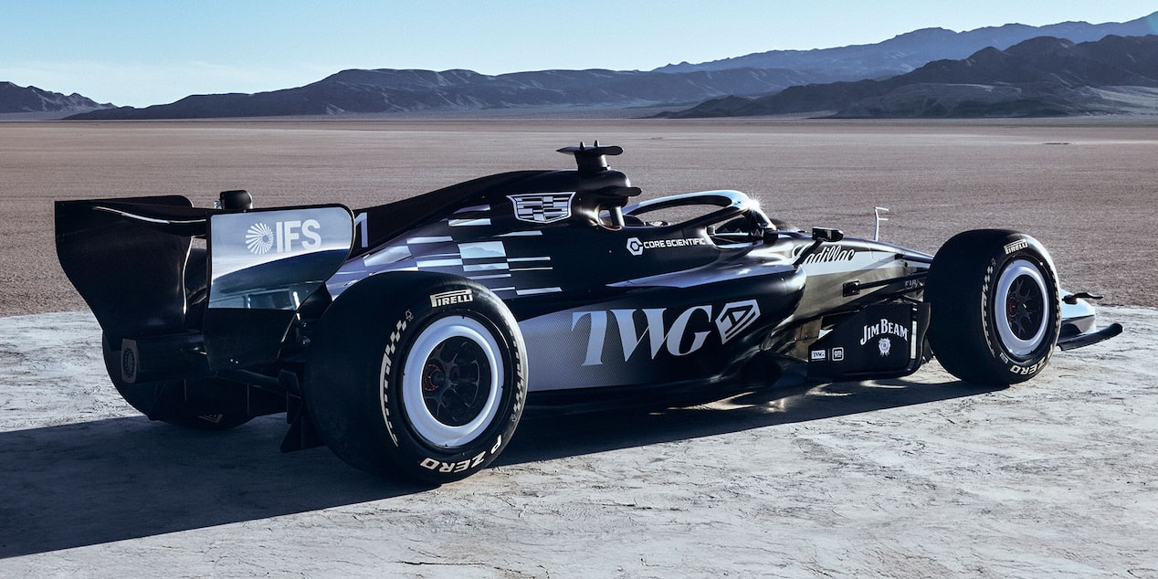 A Sleek Black-And-White Formula Race Car With Prominent Cadillac Branding Parked On A Sunlit Desert Flat With Mountains In the Distance.