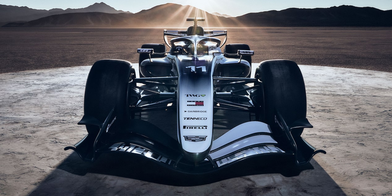 A Head-On View Of A Cadillac Formula Race Car Positioned On a Desert Road At Sunrise With Rays Of Light Casting Long Shadows Across the Scene.