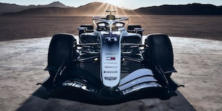 A Head-On View Of A Cadillac Formula Race Car Positioned On a Desert Road At Sunrise With Rays Of Light Casting Long Shadows Across the Scene.
