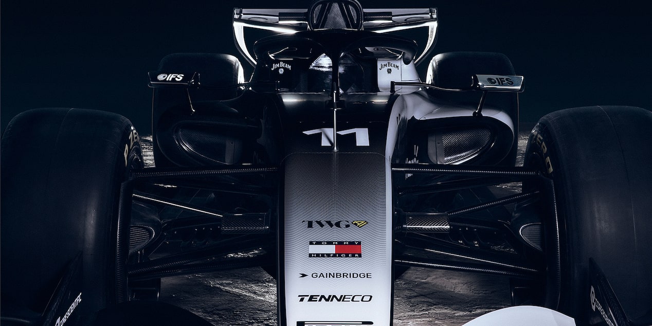 A Close Front View Of A Cadillac-Branded Formula Race Car Highlighting the Nosecone, Suspension Arms, And Aerodynamic Bodywork Against A Dark Backdrop.