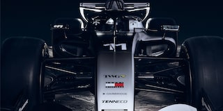 A Close Front View Of A Cadillac-Branded Formula Race Car Highlighting the Nosecone, Suspension Arms, And Aerodynamic Bodywork Against A Dark Backdrop.