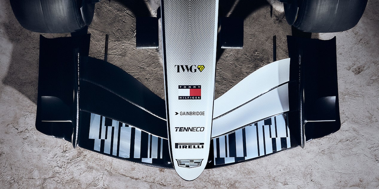 A Detailed Close-Up Of the Front Wing And Nosecone Of a Cadillac Formula Race Car Showing Aerodynamic Fins And Sponsor Markings Against a Dusty Desert Surface.