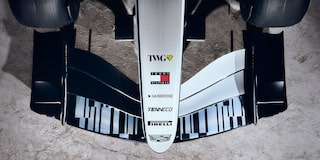 A Detailed Close-Up Of the Front Wing And Nosecone Of a Cadillac Formula Race Car Showing Aerodynamic Fins And Sponsor Markings Against a Dusty Desert Surface.