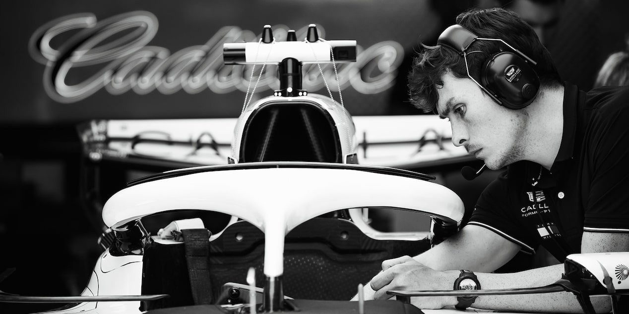 Black And White Detail View Of Engineer Working On F1 Car In Team Garage With Cadillac Logo In Background.