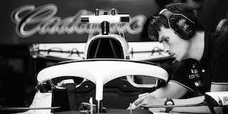 Black And White Detail View Of Engineer Working On F1 Car In Team Garage With Cadillac Logo In Background.