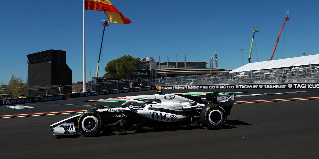 El auto de carrera Cadillac F1 de rines descubiertos recorre a gran velocidad una curva del circuito durante una sesión de entrenamientos, con una enorme bandera española ondeando al fondo.
