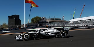 El auto de carrera Cadillac F1 de rines descubiertos recorre a gran velocidad una curva del circuito durante una sesión de entrenamientos, con una enorme bandera española ondeando al fondo.