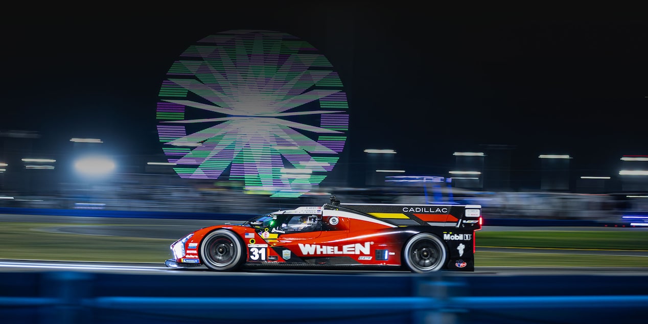 A Red Whelen Cadillac Race Car Speeding Along a Track at Night With a Large Illuminated Ferris Wheel Glowing in the Background.