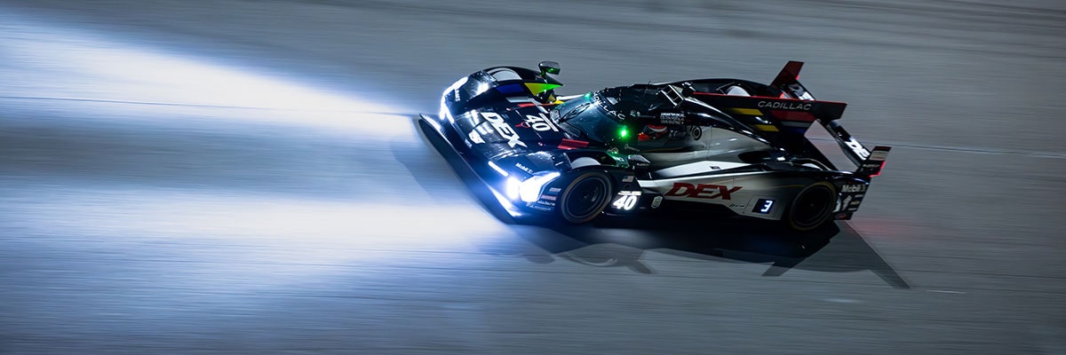 A Black Whelen Cadillac V-Series.R Race Car Drifts Through a Turn at Night With Its Headlights Casting Bright Beams Across the Track.