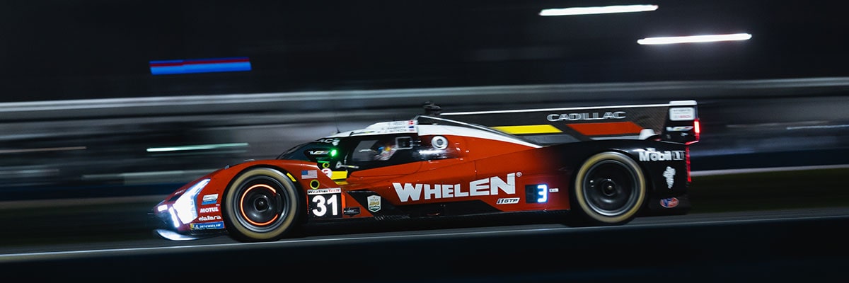 A Red Whelen Cadillac V-Series.R Race Car Speeds Down the Track at Night With Its Headlights Glowing and Motion Blur Emphasizing Its High Velocity.