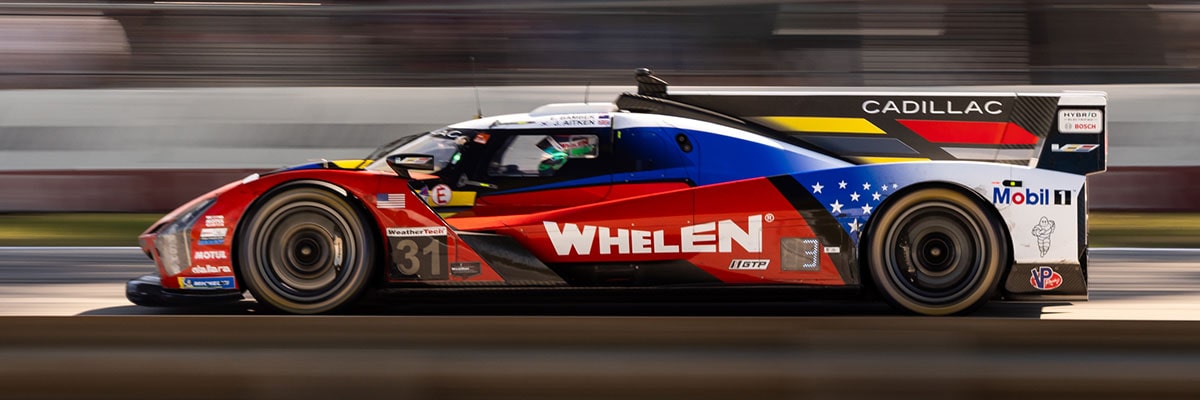 A Red Whelen Cadillac V-Series.R Race Car Speeds Down the Track at Night With Its Headlights Glowing and Motion Blur Emphasizing Its High Velocity.