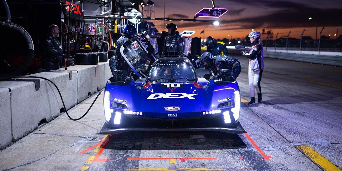 A Red Whelen Cadillac V-Series.R Race Car Races Down the Track at Dusk With Its Aerodynamic Body Low and Fast Against the Asphalt.