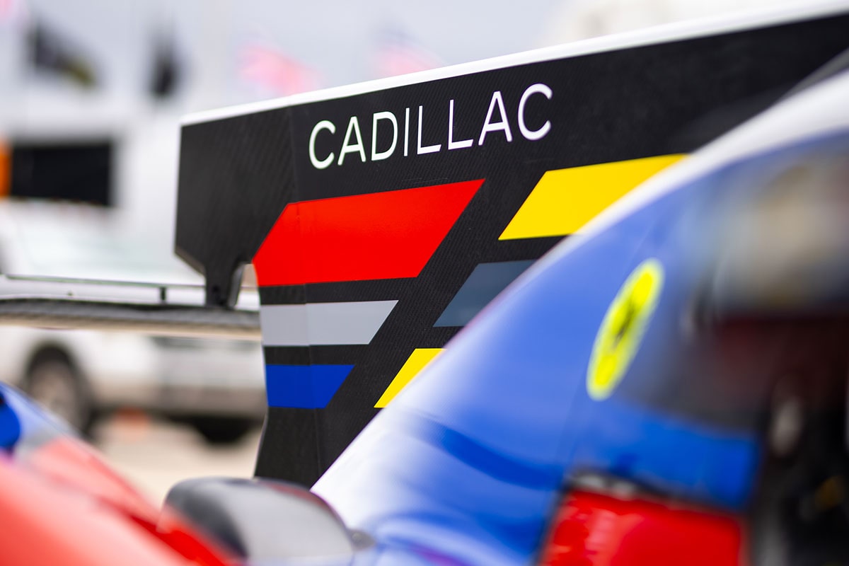 A Whelen Cadillac V-Series.R Race Car Speeds Along the Track at Night With a Large Multicolored Ferris Wheel Glowing Brightly Behind It.