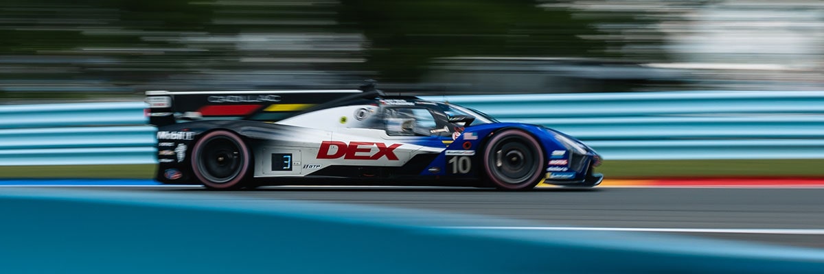 A Blue and White DXC Technology–Branded Cadillac Race Car Driving at High Speed With Motion Blur Across a Blue Track Surface.