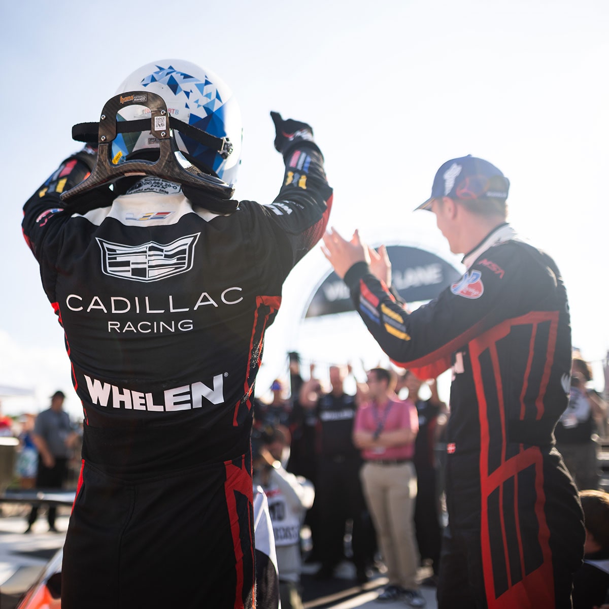 A Cadillac Racing Driver Wearing a Whelen-Branded Suit Raising Both Arms While Facing Another Team Member Along a Pit Lane With a Crowd in the Background.